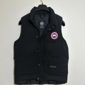 Canada Goose (pris kan diskuteras) - Svart dunväst från Canada Goose med klassisk logga på bröstet. Västen har hög krage, tryckknappar framtill och två stora fickor. Jag säljer den pågrund av att de är mitt ex och han har lämnat kvar den så jag säljer (pris kan diskuteras) snabb affär 