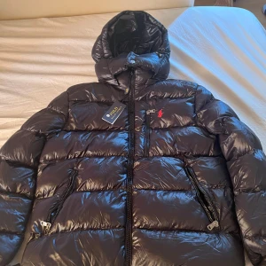 Svart dunjacka från Polo Ralph Lauren - Snygg svart dunjacka från Polo Ralph Lauren med glansig finish, huva och dragkedjor på fickorna. Jackan har klassisk pufferstil och röd broderad logga på bröstet. Perfekt för kalla dagar när du vill hålla stilen och värmen.