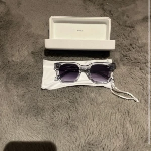 Chimi  solglasögon - Snygga solglasögon från Chimi med transparent båge och fyrkantig form. Glasen är mörktonade i grått och bågarna har en cool glittrig detalj på sidorna. Perfekta för att lyfta din stil under soliga dagar.