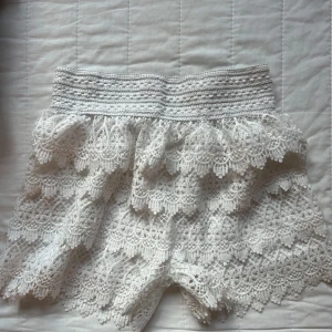 Vita spetsiga shorts med volang - Superfina vita shorts med bred resår i midjan och flera lager spets med volang. De har en romantisk och bohemisk vibe, perfekta för varma dagar. Spetsen är detaljerad och ger shortsen en unik look. Sitter skönt tack vare den stretchiga midjan.