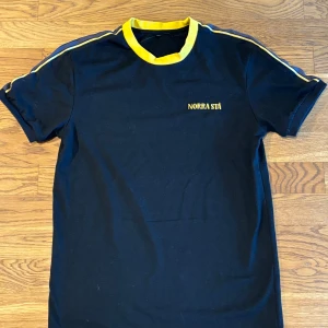 AIK t-shirt  - Svart t-shirt från Norra Stå med gul ribbad krage och gula ränder längs axlarna. Broderad gul text på bröstet. Skick 10/10 då den aldrig är använd 