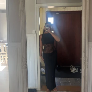 Svart långklänning med spetsdetalj - Säljer en svart långklänning med hög hals och transparent spets i midjan. Klänningen är ärmlös och har en elegant, figurnära passform. Perfekt för dig som vill sticka ut med en stilren och sofistikerad look.