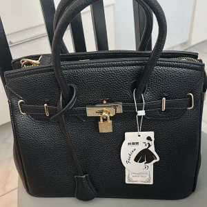 Svart handväska med gulddetaljer - Stilren svart handväska i skinnimitation med guldfärgade metalldetaljer och lås framtill. Väskan har dubbla handtag och strukturerad yta, perfekt för dig som vill ha en klassisk och snygg accessoar till din outfit.