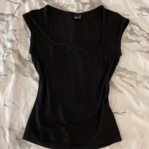 Svart topp med fyrkantig ringning Gina Tricot - En svart, tight topp från Gina Tricot med snygg fyrkantig ringning och ärmlös design. Enkel och stilren modell som sitter figurnära och passar perfekt till jeans eller kjol. Gjord i mjukt bomullsmaterial för en bekväm känsla.