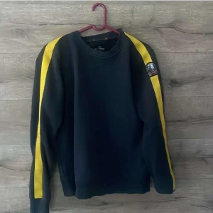 Svart fleece-tröja Parajumpers gul detalj - Svart fleece-tröja från Parajumpers med gula ränder längs ärmarna och logga på axeln. Tröjan har rund hals och lång ärm, perfekt för dig som gillar sportig och clean stil. Materialet är mjukt fleece och modellen är normal i passformen.