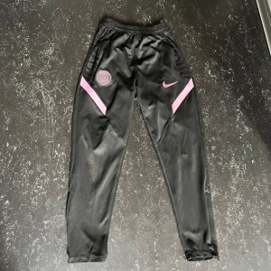 PSG träningsbyxor  - Svarta träningsbyxor från Nike med Paris Saint-Germain-logga och rosa detaljer. Byxorna har elastisk midja, smal passform och är gjorda i Dri-Fit polyester som andas. Perfekta för fotboll eller träning, med snygg rosa Nike Swoosh och PSG-märke på låret. M och passar S , skriv för mer info!!