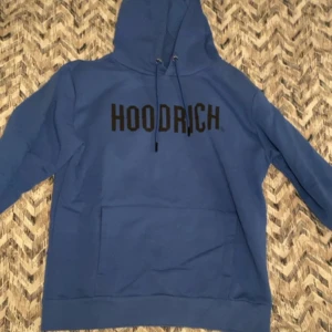Blå HOODRICH hoodie med tryck - Blå hoodie från HOODRICH med svart tryck över bröstet. Klassisk känguruficka framtill, huva med snörning och ribbade muddar. Perfekt för dig som gillar streetwear och vill ha en skön och snygg tröja.