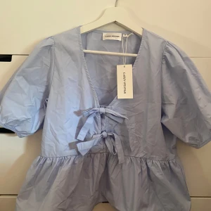 Ljusblå blus med knyt från Carin Wester - Supersöt ljusblå blus från Carin Wester med puffiga korta ärmar och två rosetter  framtill. Blusen har v-ringning och volang nedtill som ger en luftig och trendig look.  Pris kan diskuteras