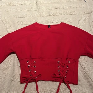 Röd croppad tröja med snörning FB Sister - Cool röd croppad tröja från FB Sister med långa ärmar och snygga snörningsdetaljer i midjan. Tröjan har ribbade muddar och är gjord i mjuk bomull, perfekt att matcha till jeans 