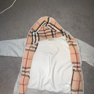 Grå hoodiejacka från Burberry M - Snygg grå hoodiejacka från Burberry med klassiskt rutigt foder i beige, svart och rött. Jackan har dragkedja, huva och två fickor framtill. Perfekt för dig som vill ha en stilren streetlook med lyxig touch. Dragkedjan funkar inte, men det går och fixa för 50-150kr!