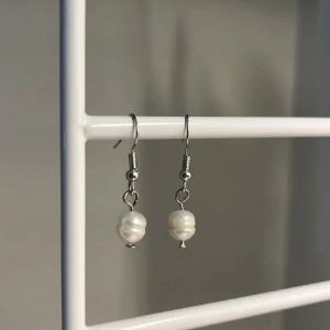 Silvriga örhängen med vita pärlor - Snygga örhängen med hängande vita pärlor och silvriga detaljer. Perfekta för att ge din look en elegant touch. Pärlorna har en naturlig, oregelbunden form som gör dem unika och trendiga. Handgjort 