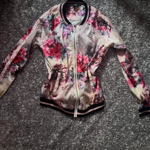 Blommig bomberjacka Sofie Schnoor - Använd ett fåtal gånger men är i bra skick. Snygg bomberjacka från Petit Sofie Schnoor med blommigt mönster i rosa, rött och grått på ljus botten. Jackan har svarta och rosa ribbade muddar, dragkedja framtill och klassisk bomberkrage. Perfekt för dig som vill sticka ut med en färgglad och trendig look.