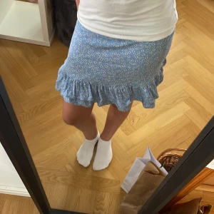 Somrig blå volang kjol - Fin blå volang kjol men blommor på. Kjolen har elastiska band där bak så den passar länge! Den är i väldigt gott skick och har inget tecken på användning!