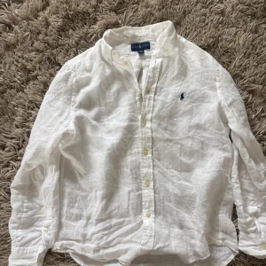 Vit linneskjorta från Ralph Lauren - Klassisk vit skjorta från Ralph Lauren i svalt linne. Skjortan har lång ärm, knappar hela vägen och den ikoniska broderade loggan på bröstet. Perfekt för en clean och stilren look.