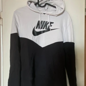 Nike svartvit hoodie med logga - Nike hoodie i svart och vitt med stor logga framtill. Tröjan har huva med snörning, lång ärm och klassisk känguruficka. Perfekt för chill dagar eller när du vill ha en sportig look. Mjuk bomullsblandning som känns skön mot huden. Frakt tillkommer