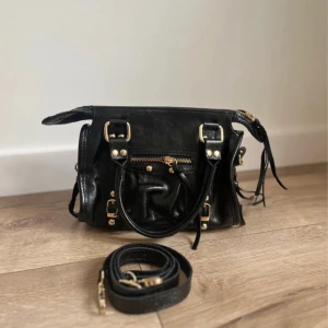 Svart handväska i skinn med gulddetaljer - Cool svart handväska i skinn med snygga guldfärgade detaljer och dragkedja upptill. Väskan har både handtag och avtagbar axelrem. 