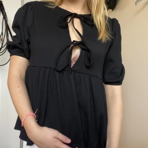 Svart blus med knyt och puffärm - Supersöt svart blus med två knyt framtill och lätt puffade korta ärmar. Modellen är luftig och har en flowy passform som ger en chill vibe. Perfekt för dig som gillar trendiga detaljer och vill sticka ut lite extra.