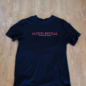 Svart t-shirt Cloud Reveal från Smog - Svart t-shirt från Smog med relaxed fit. Framsidan har trycket 'CLOUD REVEAL' i rött och baksidan har en stor örn-grafik och text. Skön bomullskvalitet och klassisk rund hals. 