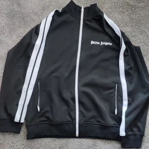 Palm Angels svart track jacket M - Palm Angels svart track jacket i storlek M med vita ränder längs ärmarna och broderad logga på bröstet. Jackan har hel dragkedja framtill, två fickor med dragkedja och ribbade muddar. Tillverkad i 100% polyester för en sportig och clean look.
