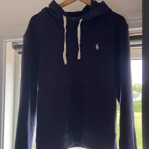 Marinblå hoodie från Polo Ralph Lauren - Säljer en marinblå hoodie från Polo Ralph Lauren med vit broderad logga på bröstet och vita snören i huvan. Klassisk känguruficka framtill och mjukt bomullsmaterial. Perfekt för dig som gillar stilren och avslappnad look.