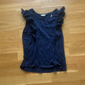 Mörkblå volangtopp Holly & Whyte - Supersöt mörkblå ärmlös topp från Holly & Whyte by Lindex. Toppen har volangdetaljer med broderade spetsar längs axlarna och sidorna, vilket ger en romantisk och trendig look. Perfekt att styla med jeans eller kjol för en cool vibe.