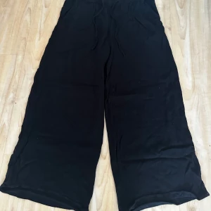 Svarta vida byxor från Indiska - Svarta byxor från Indiska i storlek S. De har en loose och wide passform med resår och snörning i midjan. Lätta och luftiga, perfekta för chill dagar eller när du vill ha något bekvämt men ändå snyggt.