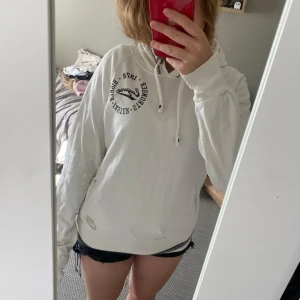 Vit hoodie från Nike med tryck - Säljer en vit hoodie från Nike med svart tryck på bröstet och broderad Nike-logga vid fickan. Tröjan har huva med snörning, långa ärmar och ribbade muddar samt randig detalj längst ner. Perfekt för chill dagar eller när du vill ha en sportig look.