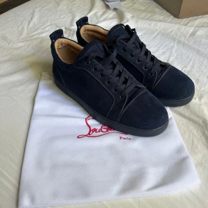 Mörkblå mocka sneakers från Louboutin - Snygga mörkblå sneakers från Christian Louboutin i mocka med klassisk röd sula och svarta snören. Skorna har rund tå, platt sula och lyxig känsla med beige skinnfoder. Perfekta för dig som vill sticka ut med exklusiva detaljer.