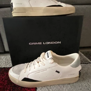 Crime London vita sneakers med mockadetaljer - Snygga vita sneakers från Crime London med beige sula, grå mockadetaljer på tån och hälen samt svarta inslag på sidan och bak. Skorna har snörning och textdetaljer på sidan. Perfekta för dig som gillar stilrena men ändå unika sneakers.