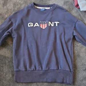 Blå sweatshirt från GANT med brodyr - Säljer en marinblå sweatshirt från GANT med broderad logga och sköld på bröstet. Tröjan har rund hals, ribbade muddar och är tillverkad i mjuk bomull. Perfekt för en chill och stilren look.