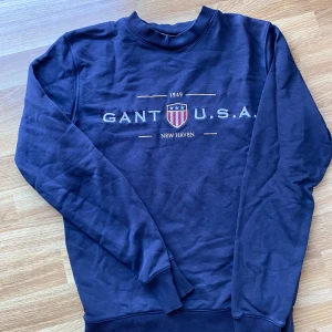 Mörkblå sweatshirt från Gant - Säljer en mörkblå sweatshirt från Gant med broderad logga och USA-sköld på bröstet. Tröjan har ribbade muddar vid hals, ärmslut och nederkant samt röd insida i kragen. 
