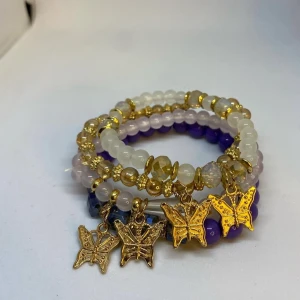 Armband med fjärilsberlocker i guld - Flera elastiska armband med pärlor i vitt, lila och guldtoner. Varje armband har detaljer i guld och är dekorerat med hängande fjärilsberlocker i guldfärg. Pärlorna är både matta och glansiga. Dessa armband är inte använda utan kommer från min small business. 