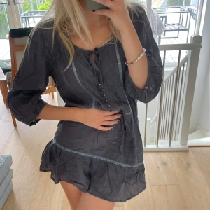 Mörkgrå kort klänning från M&Co - Supersöt mörkgrå kort klänning från M&Co med knytning i halsen, dekorativa knappar och spetsdetaljer framtill. Klänningen har trekvartsärmar och volang nertill! Supercool och snygg, vardags och fest 🖤 skulle säga att den går att användas xs-m beroende på hur den ska stylas!