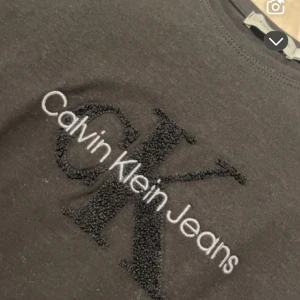 Svart Calvin Klein Jeans t-shirt - Svart t-shirt från Calvin Klein Jeans med stort broderat CK-logo och vit text på bröstet. Klassisk rund halsringning och korta ärmar. Tillverkad i mjuk bomull för en skön känsla. Perfekt för dig som gillar stilrena och trendiga plagg. Passar en xs