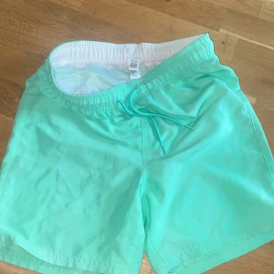 Mintgröna badshorts med snörning - Säljer ett par mintgröna badshorts med elastisk midja och snörning framtill. Shortsen har innerbyxa i mesh och är perfekta för strand eller pool. Lätta och snabbtorkande, passar dig som gillar en fräsch och enkel stil.
