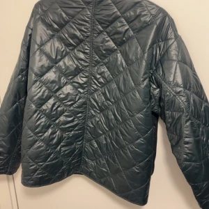 Mörkgrön quiltad jacka från Shinamoo - Snygg mörkgrön quiltad jacka från Shinamoo med lång ärm och V-ringad hals. Jackan har ett stilrent, minimalistiskt utseende utan synliga knappar eller dragkedja. Perfekt för lager-på-lager under hösten eller våren.