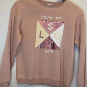 Rosa sweatshirt med paljetter och text - Säljer en ljusrosa sweatshirt med lång ärm och rund hals. Framsidan har en fyrkantig paljettdekor som kan vändas och bildar olika färger och texten 'YOU'RE MY SOULMATE'. Perfekt för dig som gillar unika detaljer!