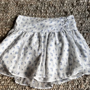 Vit blommig kjol från Gina Tricot - Supersöt vit kjol från Gina Tricot med blått blommönster och lätt volang. Kjolen har en bred midja och är gjord i ett tunt, luftigt material som känns perfekt för varmare dagar.