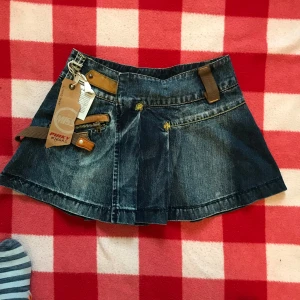 Jeanskjol med detaljer - Cool kort jeanskjol med bruna läderdetaljer och dragkedjor. Kjolen har markerade sömmar och en unik design med flera spännande detaljer framtill. Perfekt för dig som vill sticka ut med din stil.