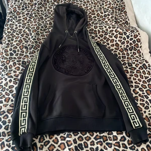 Svart hoodie från Glorious Gangsta med mönstrade band - Säljer en svart,guldig hoodie från Glorious Gangsta med stora broderade detaljer på bröstet och breda, mönstrade band längs ärmarna. Tröjan har huva med dragsko och en stor magficka. Materialet är en mix av bomull och polyester.