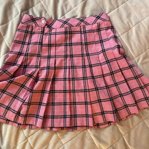 Rosa rutig kortkjol - Säljer en rosa kortkjol med svart och vit rutigt mönster. Kjolen har dragkedja och knapp i midjan samt veckad nederdel för extra volym. Perfekt för dig som gillar en färgglad och trendig stil.