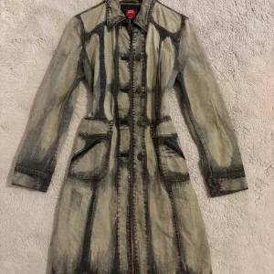 Trench coat från Miss Sixty - Säljer en lång, figursydd jeansjacka från Miss Sixty med unikt tvättad denimlook. Jackan har knappar framtill, stora fickor med dragkedja och markerade sömmar. Snygga detaljer som förstärkta armbågar och klassisk krage.