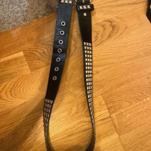 Svart nitar bälte - Säljer ett svart bälte med silvriga nitar i metall. Bältet har flera hål för justering och ett coolt, punkigt utseende. Perfekt accessoar för att ge outfiten edge.
