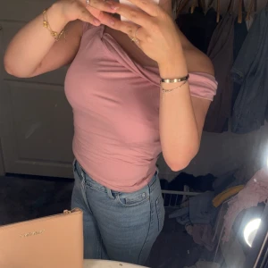 Rosa offshoulder topp - Säljer en ljusrosa offshoulder topp med draperad halsringning. Toppen är tight och har en snygg, asymmetrisk axel. Perfekt för att lyfta en enkel outfit med en trendig touch.