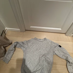 Grå sweatshirt från Stone Island - Säljer en grå sweatshirt från Stone Island med klassisk logotyp-patch på ärmen. Tröjan har rund halsringning och långa ärmar. Perfekt för en avslappnad och stilren look.
