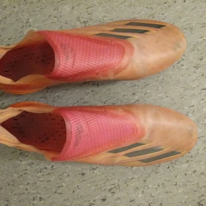 Adidas fotbollsskor i orange och rosa - Säljer ett par Adidas fotbollsskor i en snygg orange och rosa färgkombination. Skorna har en strömlinjeformad design utan snörning och tydliga svarta Adidas-ränder på sidan. Perfekta för dig som vill sticka ut på planen!