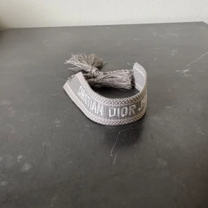 Grått armband från Dior med tofsar - Säljer ett snyggt grått armband från Dior med broderad logga och dekorativa tofsar i ändarna. Perfekt accessoar för att lyfta din outfit med en exklusiv touch.