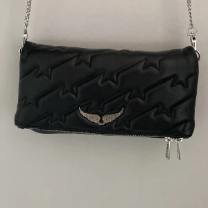 Zadig & Voltaire Väska - Cool svart axelväska med quiltat blixtmönster och silverfärgad kedjerem. Väskan har dragkedja både fram och bak samt en snygg metallvinge på framsidan. Perfekt för att lyfta din outfit med en edgy touch.