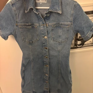 Jeansklänning med kort ärm från Zara - Säljer en klassisk jeansklänning från Zara i ljusblå denim. Klänningen har korta ärmar, knappar framtill och två bröstfickor. Perfekt för en avslappnad och trendig look.