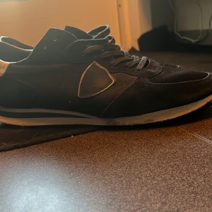 Svarta Philippe modell trainers - Säljer ett par svarta sneakers från Philippe Model med vita detaljer på hälen och sidorna. Skorna har snörning, rund tå och platt sula. Perfekta för en stilren och sportig look.